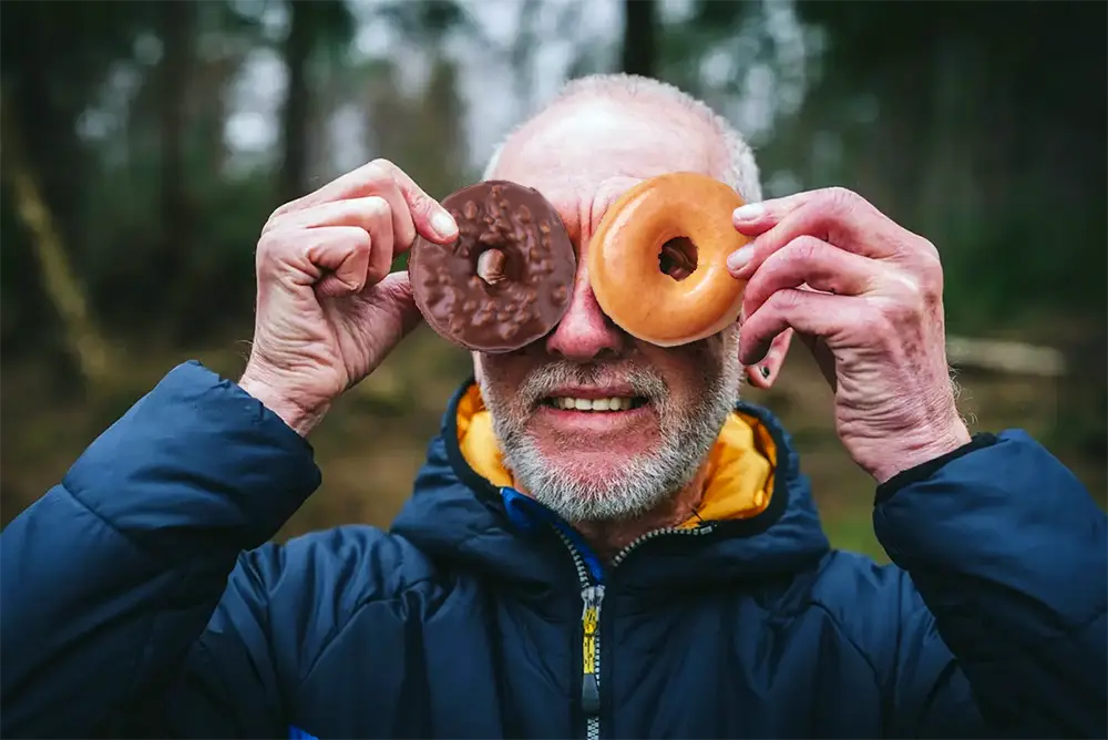 Abuelo sonriente sosteniendo una dona de chocolate y una dona glaseada de Krispy Kreme.