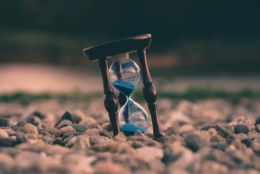 Un reloj de arena de madera con arena azul inclinado sobre piedras, simbolizando la percepción subjetiva del tiempo y la sensación de lentitud en ciertos momentos.