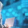 Hacker con capucha usando laptop frente a código binario en pantalla azul.