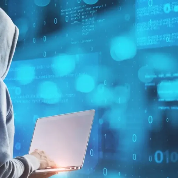 Hacker con capucha usando laptop frente a código binario en pantalla azul.