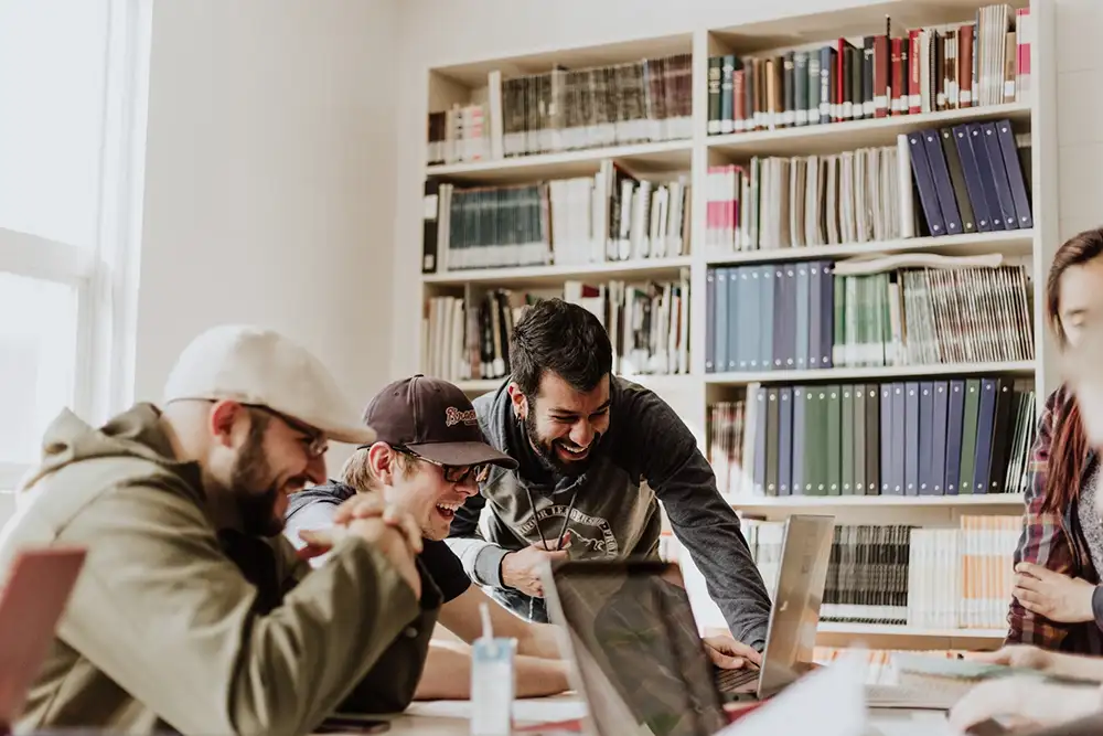 Estudiantes colaborando en biblioteca con laptops, desarrollando habilidades de comunicación y trabajo en equipo.