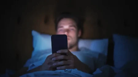 Hombre acostado en la cama usando su teléfono móvil en la oscuridad, iluminado por la pantalla del dispositivo.