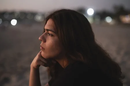 Retrato en primer plano de una mujer joven con expresión contemplativa en la playa durante el anochecer, con luces desenfocadas en el fondo.