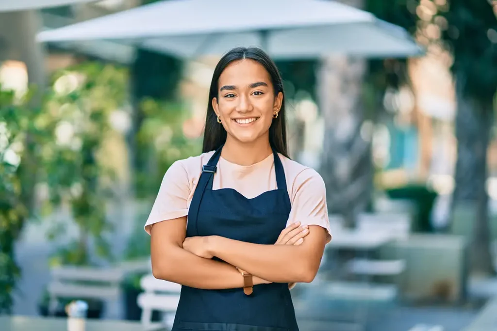 Mujer emprendedora con delantal sonriendo en un café al aire libre, simbolizando el acceso a oportunidades financieras.