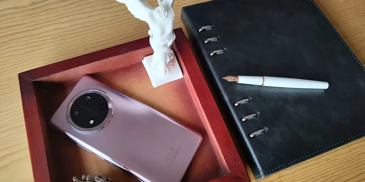 HONOR Magic 7 Lite en color Titanium Purple junto a una pluma estilográfica blanca, agenda negra, escultura blanca y decoración sobre mesa de madera, destacando diseño elegante con módulo de cámara circular.