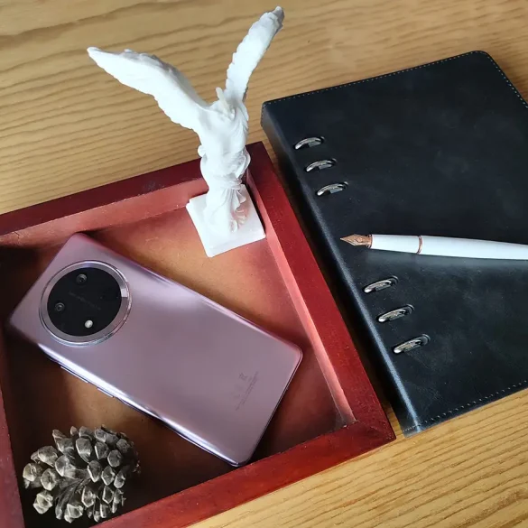 HONOR Magic 7 Lite en color Titanium Purple junto a una pluma estilográfica blanca, agenda negra, escultura blanca y decoración sobre mesa de madera, destacando diseño elegante con módulo de cámara circular.