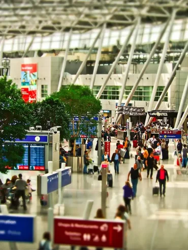 Terminal de aeropuerto internacional con viajeros caminando, señalizaciones de vuelos, y áreas de espera bajo una estructura moderna con techos altos.