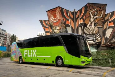 Autobús verde de Flix estacionado frente al emblemático mural del Polyforum Cultural Siqueiros en Ciudad de México, marcando el inicio de operaciones de la empresa alemana en el país.