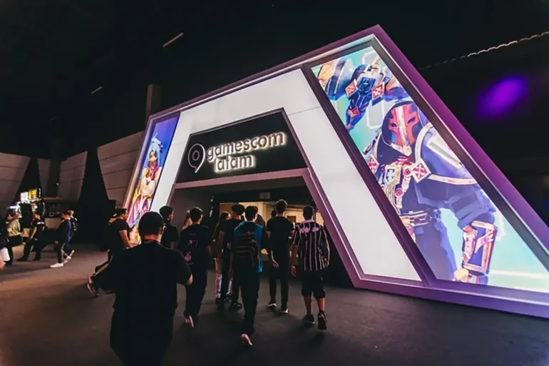 Entrada principal de gamescom latam 2025 en São Paulo, con asistentes ingresando al recinto bajo una estructura iluminada y pantallas LED con arte de videojuegos.