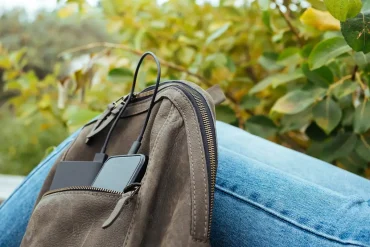 Mochila tecnológica con dispositivo móvil y batería externa conectados por cables USB, apoyada sobre una pierna con jeans, en un entorno natural con vegetación al fondo.