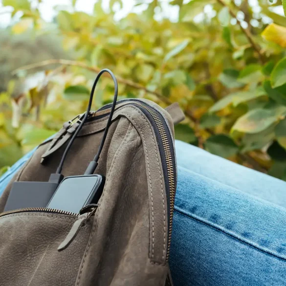 Mochila tecnológica con dispositivo móvil y batería externa conectados por cables USB, apoyada sobre una pierna con jeans, en un entorno natural con vegetación al fondo.