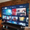 Smart TV TCL QM6K mostrando la interfaz de Google TV en una sala moderna con muro de ladrillo y lámpara de pie, destacando películas populares y recientes en pantalla.