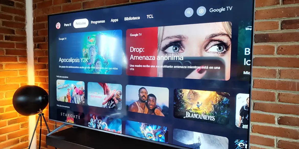 Smart TV TCL QM6K mostrando la interfaz de Google TV en una sala moderna con muro de ladrillo y lámpara de pie, destacando películas populares y recientes en pantalla.