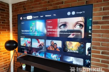 Smart TV TCL QM6K mostrando la interfaz de Google TV en una sala moderna con muro de ladrillo y lámpara de pie, destacando películas populares y recientes en pantalla.