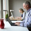 Hombre mayor sonriendo mientras usa una laptop en un espacio de trabajo compartido, con una mujer también trabajando al fondo.
