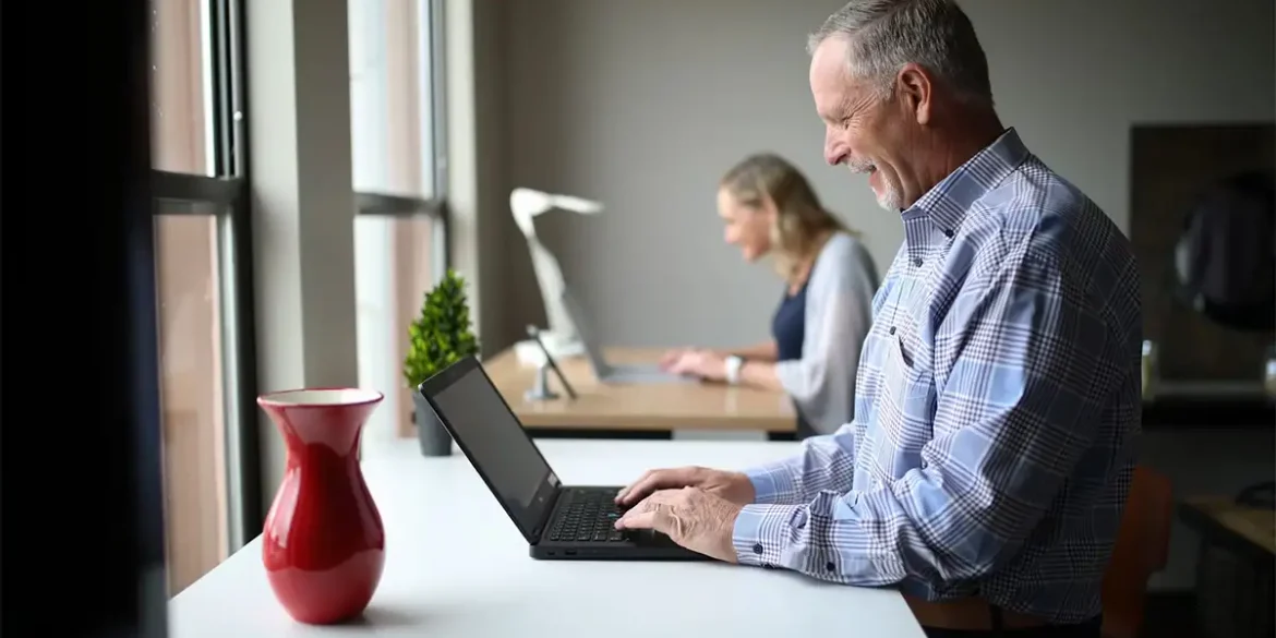 Hombre mayor sonriendo mientras usa una laptop en un espacio de trabajo compartido, con una mujer también trabajando al fondo.