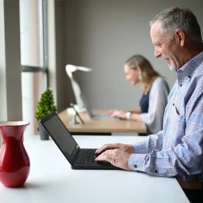Hombre mayor sonriendo mientras usa una laptop en un espacio de trabajo compartido, con una mujer también trabajando al fondo.