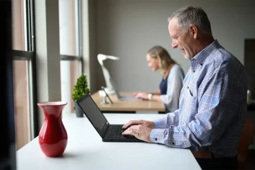 Hombre mayor sonriendo mientras usa una laptop en un espacio de trabajo compartido, con una mujer también trabajando al fondo.