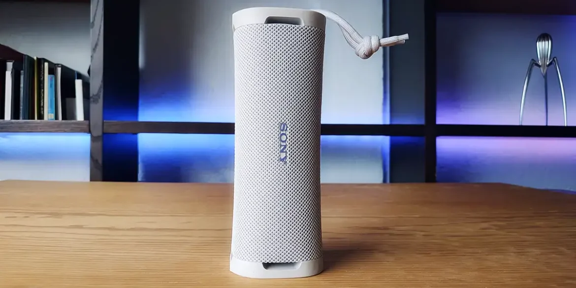 Bocina portátil Sony ULT Field 1 en color blanco, colocada en posición vertical sobre una mesa de madera, con fondo iluminado en tonos azules y morados, ideal para interiores modernos. Destaca su diseño compacto, textura de malla y correa ajustable.