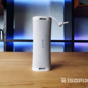 Bocina portátil Sony ULT Field 1 en color blanco, colocada en posición vertical sobre una mesa de madera, con fondo iluminado en tonos azules y morados, ideal para interiores modernos. Destaca su diseño compacto, textura de malla y correa ajustable.