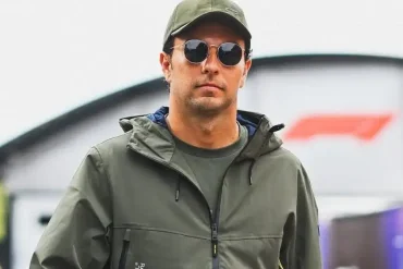 Checo Pérez camina por el paddock de la Fórmula 1 con gafas oscuras, gorra y chaqueta verde, previo al posible anuncio con Cadillac F1 en 2026.