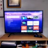 Televisor encendido con interfaz de Roku mostrando aplicaciones como Netflix, YouTube, CNET y UFC. La pantalla resalta el menú principal y apps destacadas, acompañado de su control remoto sobre un mueble de madera con objetos de uso cotidiano.