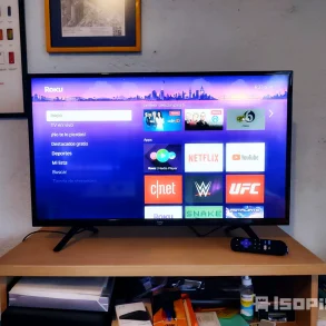 Televisor encendido con interfaz de Roku mostrando aplicaciones como Netflix, YouTube, CNET y UFC. La pantalla resalta el menú principal y apps destacadas, acompañado de su control remoto sobre un mueble de madera con objetos de uso cotidiano.