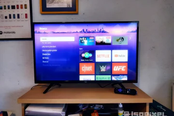 Televisor encendido con interfaz de Roku mostrando aplicaciones como Netflix, YouTube, CNET y UFC. La pantalla resalta el menú principal y apps destacadas, acompañado de su control remoto sobre un mueble de madera con objetos de uso cotidiano.