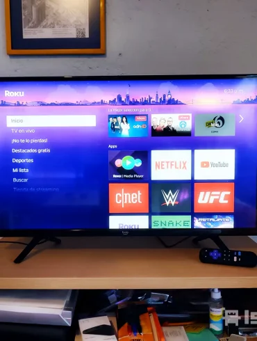 Televisor encendido con interfaz de Roku mostrando aplicaciones como Netflix, YouTube, CNET y UFC. La pantalla resalta el menú principal y apps destacadas, acompañado de su control remoto sobre un mueble de madera con objetos de uso cotidiano.