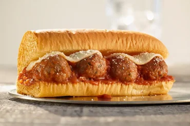 Primer plano de un Sub de Albóndigas de Subway servido en pan recién horneado, con albóndigas de res cubiertas de salsa marinara, queso mozzarella derretido y un toque de parmesano rallado, capturado sobre una superficie de madera con iluminación natural.