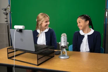 Dos estudiantes sonríen mientras participan en una videollamada educativa, utilizando una laptop Dell con cámara web y micrófono profesional, en un entorno con pantalla verde.
