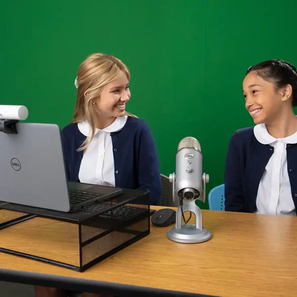 Dos estudiantes sonríen mientras participan en una videollamada educativa, utilizando una laptop Dell con cámara web y micrófono profesional, en un entorno con pantalla verde.
