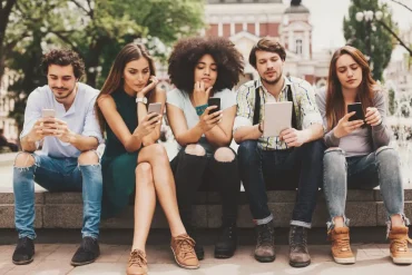 Cinco jóvenes sentados en una banca al aire libre, todos concentrados en sus dispositivos móviles y tabletas. La imagen representa el comportamiento digital de la generación actual en espacios públicos, con un enfoque en el uso intensivo de la tecnología móvil.