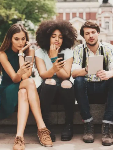 Cinco jóvenes sentados en una banca al aire libre, todos concentrados en sus dispositivos móviles y tabletas. La imagen representa el comportamiento digital de la generación actual en espacios públicos, con un enfoque en el uso intensivo de la tecnología móvil.
