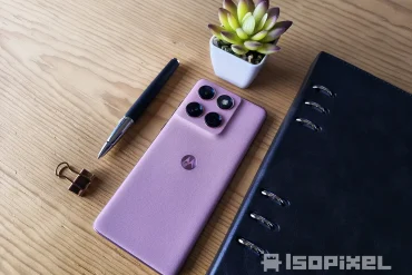 Smartphone Motorola rosa con cuatro cámaras traseras sobre mesa de madera, acompañado de pluma, clip dorado, libreta negra y maceta decorativa.