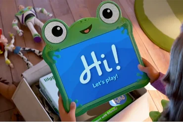 Niña sostiene tablet interactiva de rana con mensaje “Hi! Let’s play!” junto a juguetes de Toy Story en el suelo