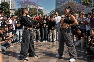 Dos mujeres compiten en una batalla de farmear aura mientras una multitud las rodea y graba con sus smartphones.
